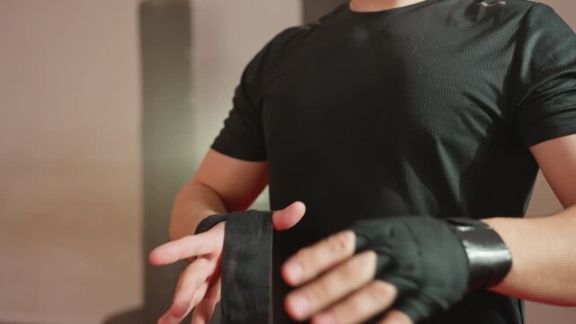 Athlete wearing black sportswear wraps hands with protective black bandages inside gym, preparing for boxing training session, showing strength, discipline, focus, and commitment before workout