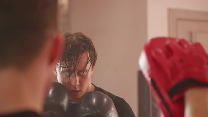 Focused boxer wearing black gloves trains with coach using red pads during intense sparring session, sweat dripping from face, eyes locked on target, demonstrating endurance, discipline in boxing gym - Powered by Adobe