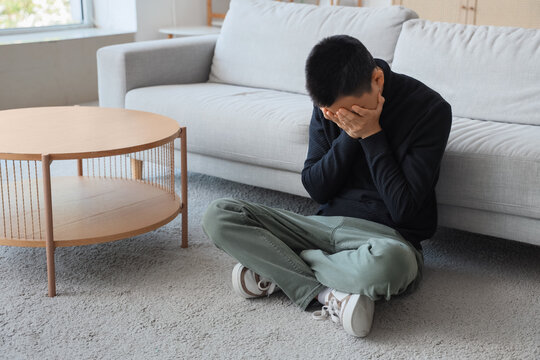 Sad young Asian man sitting on carpet at home