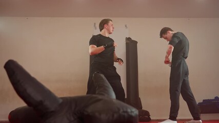 Wrestlers in gym, one athlete adjusting waistband while another lift him up and throw on red mat with punching bags and equipment visible in background