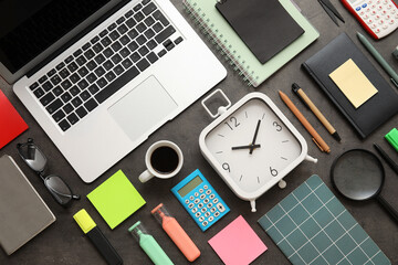 Alarm clock with laptop, cup of coffee and office supplies on dark grunge background. Time...