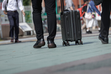 Business traveler pulls dark ribbed hardshell suitcase across busy city plaza among commuters, capturing candid everyday moment of urban movement and transit. Dutch tilt adds dynamism to scene. © thomasgweber
