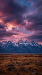 Obraz premium Dramatic sunset hues paint a vibrant sky above a snow-capped mountain range, casting a warm glow on the autumnal foreground plains