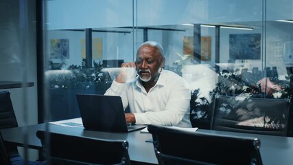 A mature African American man in a white shirt communicates via a laptop during a conference call in a modern office setting. Plants and artwork enhance the corporate environment. - Powered by Adobe