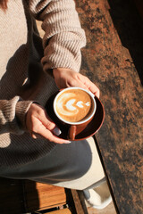 Hand holding hot coffee with latte art on wooden table under sunlight. Autumn Morning breakfast. Creamy coffee cup aromatic.