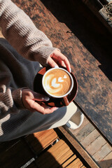 Hand holding hot coffee with latte art on wooden table under sunlight. Autumn Morning breakfast. Creamy coffee cup aromatic.