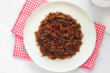 A plate of shredded smoked Skipjack Tuna with chili and spices (Cakalang Fufu Rabe), a spicy and flavorful Manado delicacy, served on a white plate with a checkered napkin.