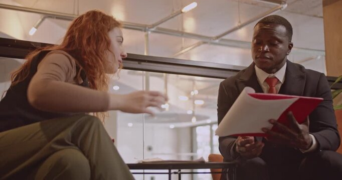 Two diverse business people discuss paperwork. A suited man holds a folder and talks with his female colleague. They're both seated in a modern office.