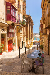Colorful traditional wooden balconies on a narrow stone staircase street in Valletta Malta with empty cafe tables in the foreground