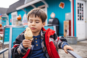 Little boy in a red winter jacket happily eating ice cream outside a colorful cafe