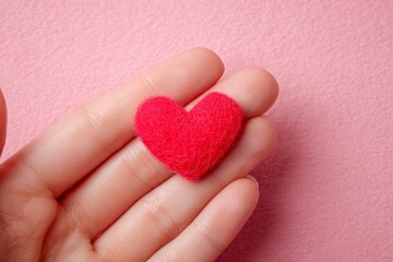 Fototapeta premium A delicate, red felt heart rests gently in a hand against a soft pink backdrop.
