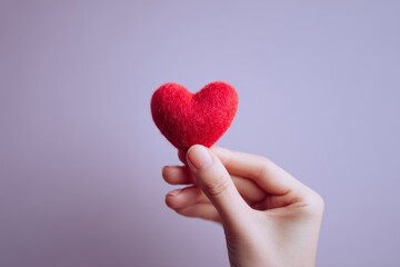 Fototapeta premium A delicate red heart, crafted from felt, is held gently in a hand against a soft lavender backdrop.