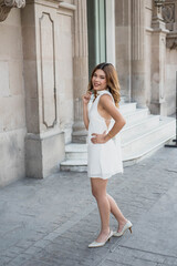 Stylish young woman in white dress posing confidently in front of modern building, radiating effortless style and sophistication with graceful elegance