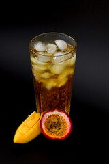 Mix of tropical fruit juices mango and passion fruit in a tall glass with ice on a black background, pieces of ripe fruit nearby.