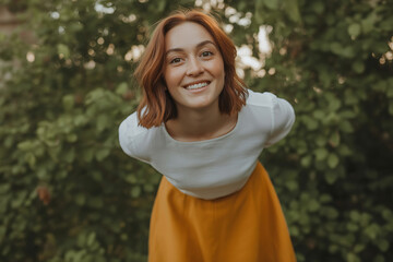 Happy redhead woman in white top and orange skirt laughing outdoors with blurred green foliage background
