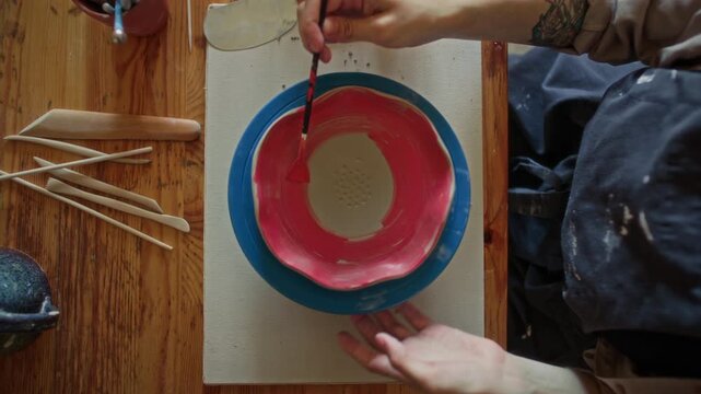 Hands of female artisan applying red glaze with paintbrush to inner surface of wavy ceramic bowl placed on rotating stand in pottery studio with carving tools nearby. Top down view