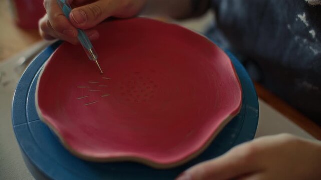 Hands of ceramic artist using scribe tool to engrave thin decorative lines on red glazed plate placed on turntable in pottery studio. Close-up view