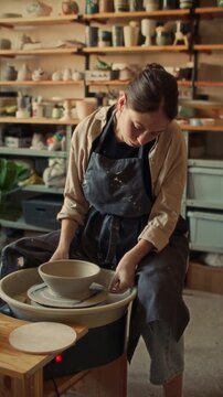 Young female artisan carefully lifting finished clay bowl from pottery wheel and placing it on plaster bat in ceramics workshop. Vertical clip