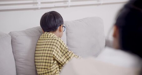 Asian boy with adhd sits cross-legged on sofa during evaluation by female psychologist, holding toy and avoiding eye contact, indicating emotional tension and challenges in behavior adjustment