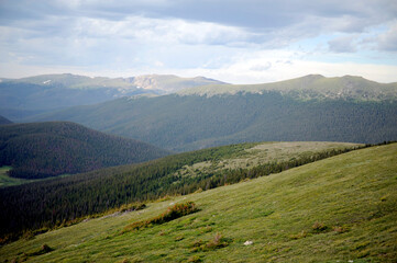 Naklejka premium Majestic mountain landscape in the morning with clouds over green grass and a vast sky, offering an alpine view of peaks and forest