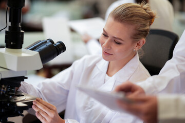 A focused female science student in a white lab coat carefully adjusts a microscope, examining a specimen during a practical lesson or research project in a university laboratory.