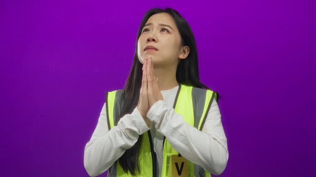 Chinese woman in reflective vest stands in a prayerful pose against a vibrant purple background, suggesting volunteer work and introspection.