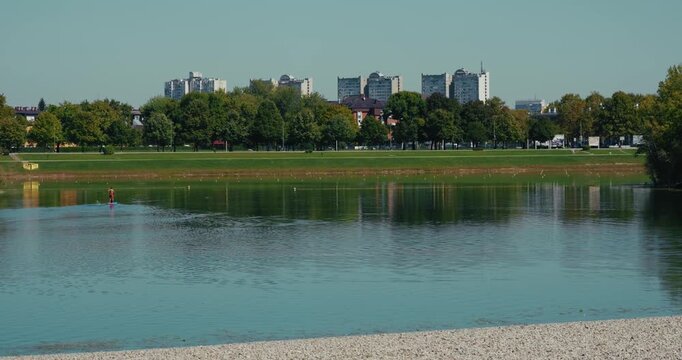 A paddleboarder moves across the calm surface of Jarun Lake with reflections of trees and residential buildings of Zagreb in the background on a clear day.