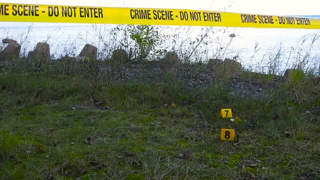 Slow tracking over crime scene tape stretched across harbor-side coastal area, fluttering in wind on daytime. Evidence markers placed beside bullets and Bitcoins scattered on the grassy seaside ground