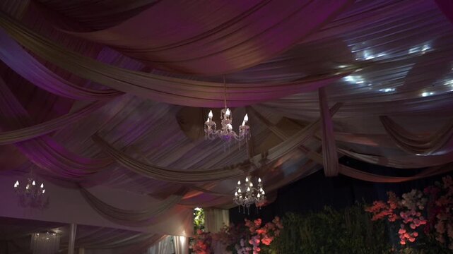 Elegant ceiling decoration with flowing fabric and glowing chandeliers under moving colorful lighting, creating a luxurious event atmosphere.