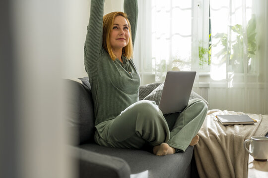 Woman with laptop sitting on couch stretching, bright room