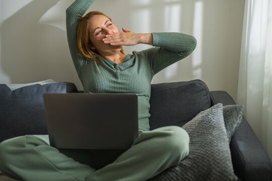 Woman with laptop sitting on couch yawning, work from home