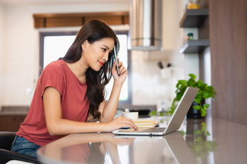 Obraz premium Young woman sitting in kitchen and working on laptop in weekend morning. Female using laptop on dining table .