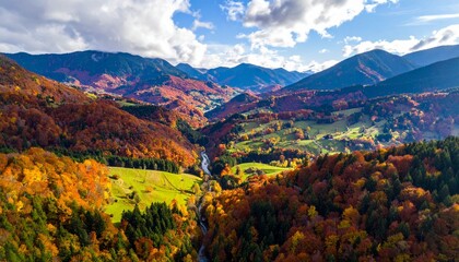 Aerial drone view of a vast mountain valley covered in vibrant, colorful autumn forests under a partly cloudy sky.