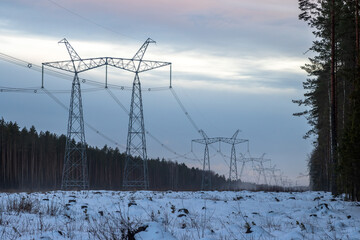 High voltage power line. View of large metal electrical poles. Power lines on a snowy clearing in the forest. Electricity supply and electric power industry. Evening industrial landscape.