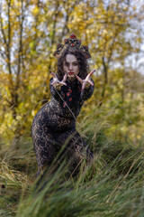 A young woman dressed as a witch and sorceress stands in an autumn forest, wearing a black dress and crown. A beautiful Gothic queen works her magic on Halloween.