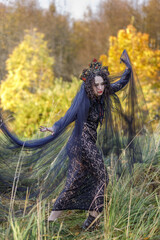A young woman dressed as a witch and sorceress stands in an autumn forest, wearing a black dress and crown. A beautiful Gothic queen works her magic on Halloween.