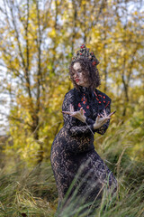 A young woman dressed as a witch and sorceress stands in an autumn forest, wearing a black dress and crown. A beautiful Gothic queen works her magic on Halloween.