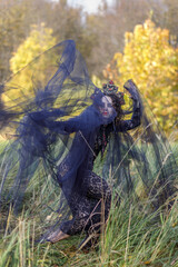 A young woman dressed as a witch and sorceress stands in an autumn forest, wearing a black dress and crown. A beautiful Gothic queen works her magic on Halloween.