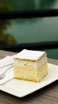 Bled cream cake Krem&scaron;nita sits on a cafe plate with Lake Bled beyond, gentle right vertical orbit.