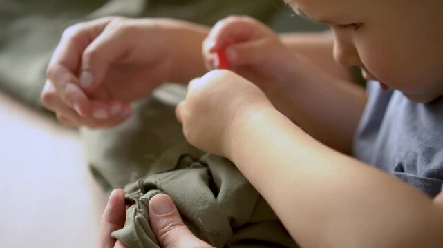 Hands of young mom repairing hem with needle while baby looks curiously. Everyday motherhood, sewing skill, repair work, shared moment of care, child eager to participate in activity.