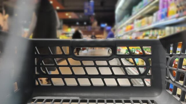 First-person view inside empty black plastic shopping cart moving down a busy supermarket aisle, blurred shelves and customers in background with bright interior lighting creating a bokeh effect.