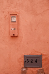 A close-up view of a weathered brown stucco wall in Morocco, featuring a metal house number plate beside a water meter. The aged textures, peeling paint, and simple utility objects
