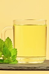 Vertical warm herbal tea in a clear glass mug with fresh mint leaves on a stone coaster, set against a studio yellow background. Refreshing wellness beverage concept, stress relief drink 