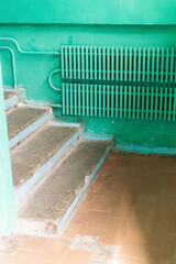 The interior of an old house reveals worn-out steps leading to a bright green wall. A rusty radiator adds to the neglected atmosphere. Dust and marks indicate years of disrepair