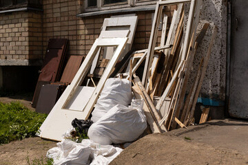 A collection of old doors and wooden scraps lies on the ground beside a brick wall. White bags of...