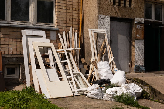 Discarded doors and construction materials are piled near a rundown building. Overgrown grass and debris hint at the neglect of this urban location during the day