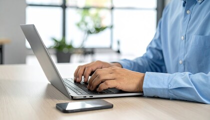 Man Typing On Laptop Computer At Desk With Smartphone Nearby In Modern Office With Plants And Natural Light