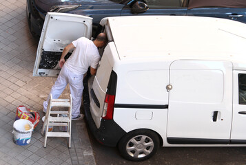 Furgoneta blanca de pintor de obra sacando herramientas del maletero con escalera, cubo y material de reforma para pintar casa