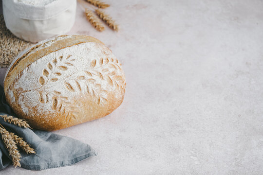 A round loaf of sourdough bread features intricate designs on its crust. It rests on a light countertop with wheat stalks and flour in the background, creating a warm, inviting scene. side view