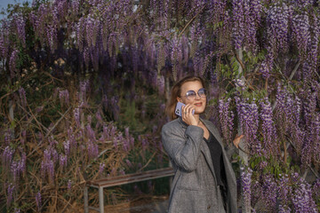 Woman Wisteria Phone Call Spring - A woman wearing a gray blazer stands in front of a flowering...